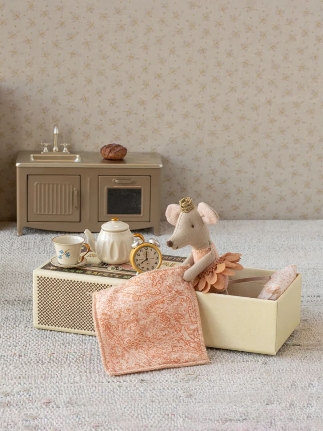 Miniature toy mouse with a blanket in a box, surrounded by miniature furniture and items on a textured surface.