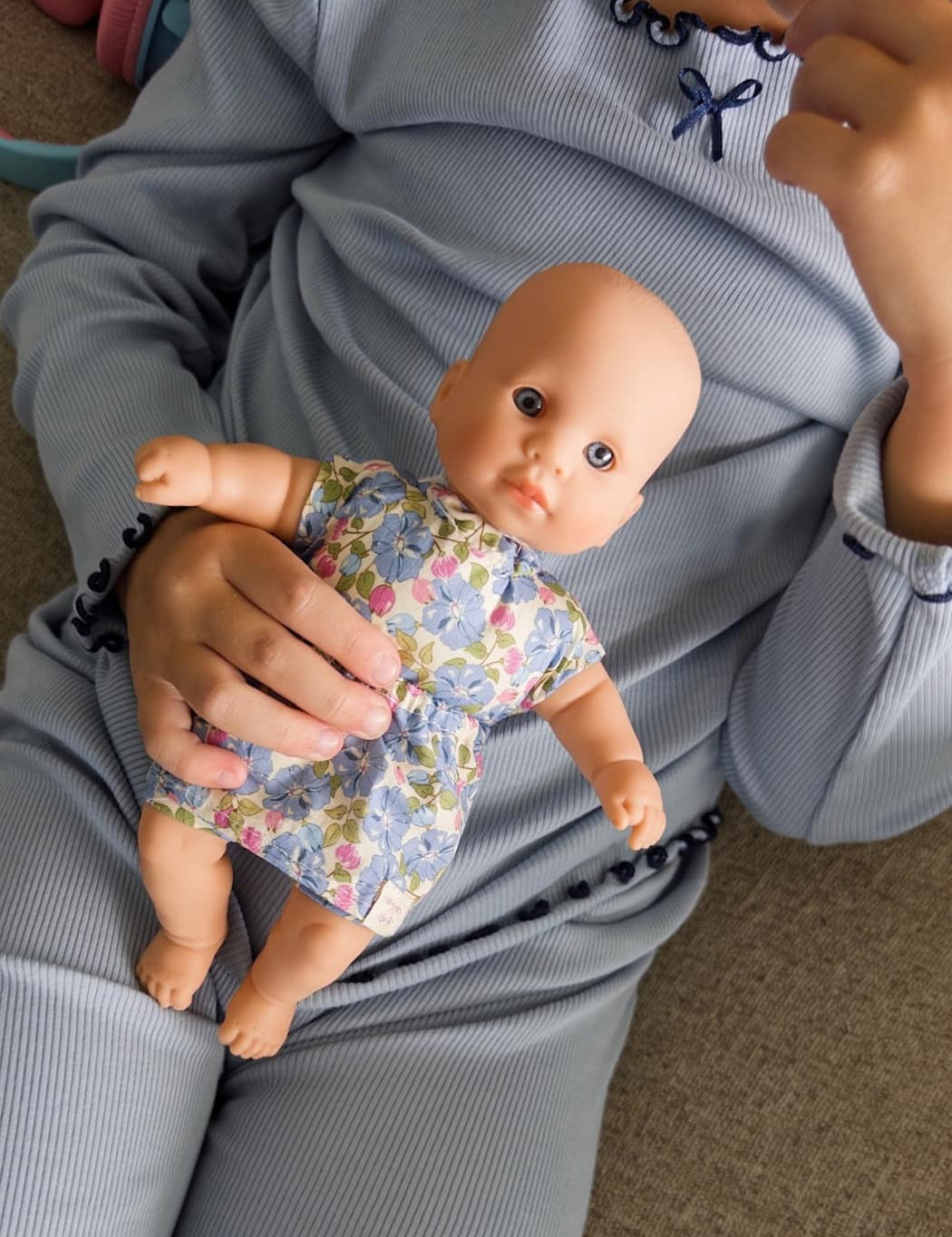 Person holding a baby doll wearing a floral outfit on a carpeted floor.