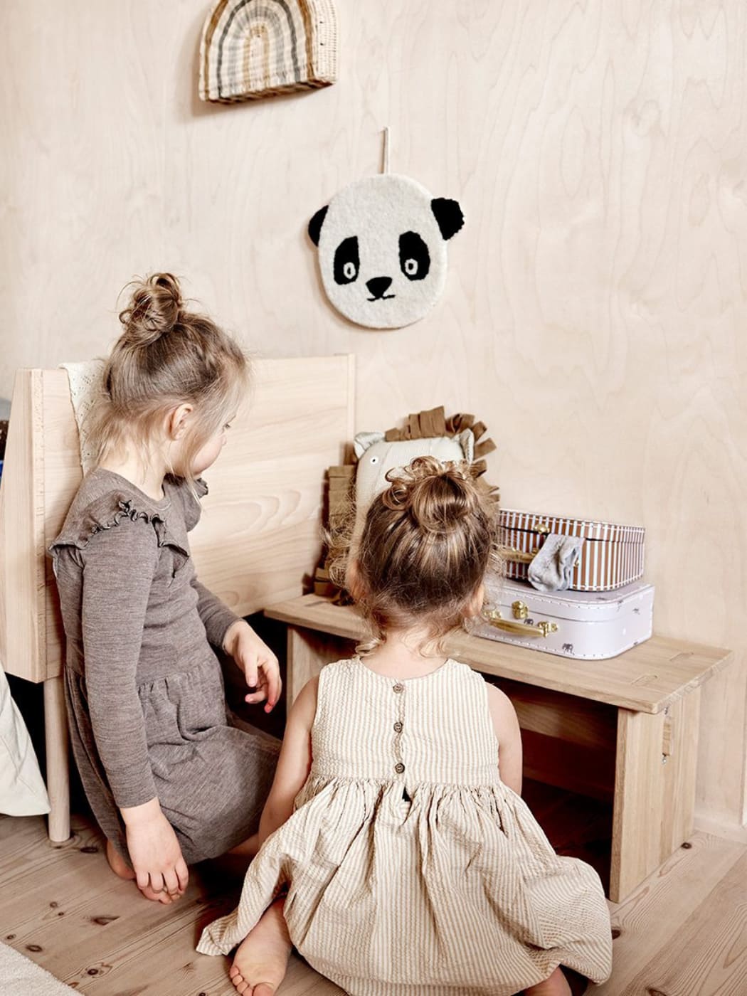 Two children sitting on a wooden floor with a panda plush toy hanging on the wall.