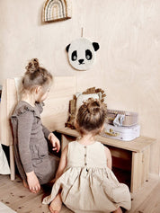 Two children sitting on a wooden floor with a panda plush toy hanging on the wall.